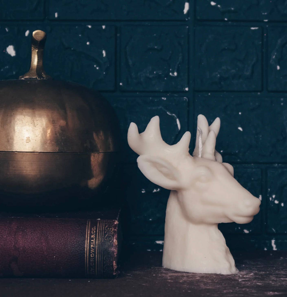 White deer head candle next to a brass object and book on a dark surface with a blue textured background