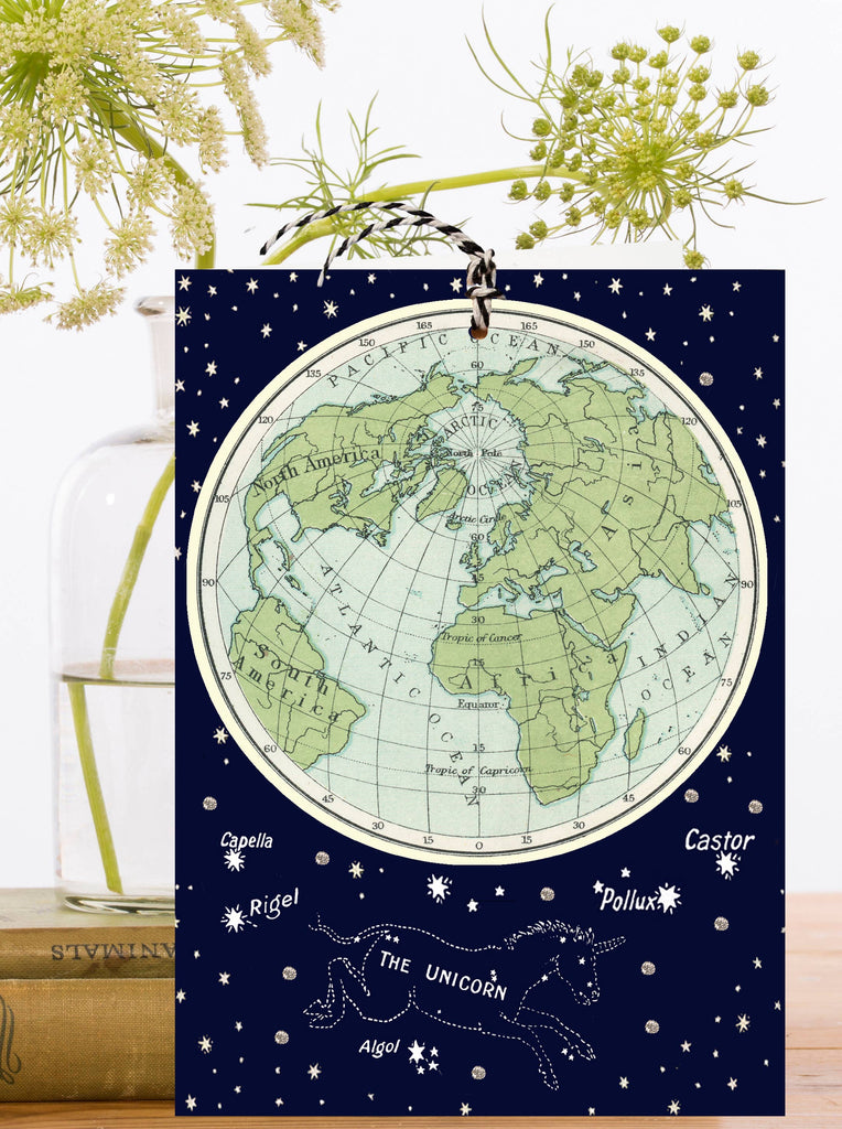 Greeting card with detachable world ornament with star constellations on a dark blue background, placed on a surface with plants and books.