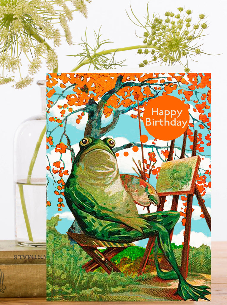Birthday card with a frog sitting in a chair painting and with flowers in the background. "Happy Birthday" text in orange circle.