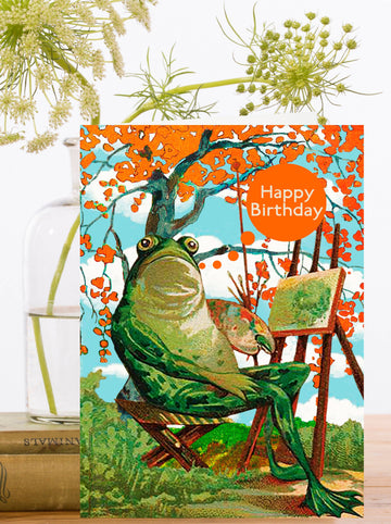 Birthday card with a frog sitting in a chair painting and with flowers in the background. "Happy Birthday" text in orange circle.