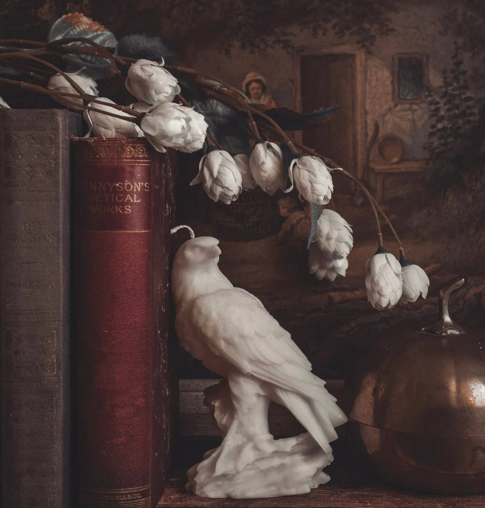 Still life with a white bird candle, white flowers, and books in a rustic setting.