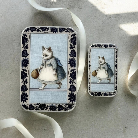 Two decorative tins with illustrations of cats in dresses on a textured surface.