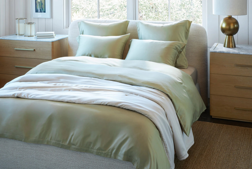 Sage green colour duvet cover and shams in softly draping Lyocell fabric with reflective sheen. In modern bedroom with large windows behind bed.