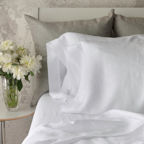 White bedding set with a pillowcase and rumpled duvet cover on a bed, styled with two gray pillows and a glass vase of white flowers on a bedside table.