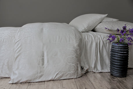 Neatly made bed with light-colored bedding with a tone on tone design and pillows against a gray wall.