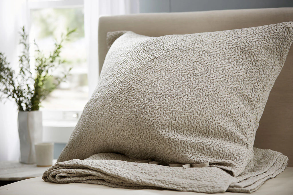 Textured beige pillow and blanket on a bed with a blurred indoor background