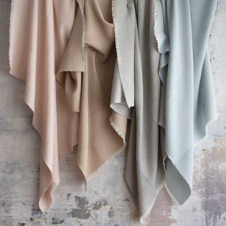 Four textured throws in dusty pink, warm beige, grey and pale blue-gray hanging side by side against a distressed gray rustic background.