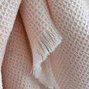 A close-up of a textured waffle-weave throw blanket in a soft blush pink color, featuring fringed edges.