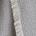 A close-up of a textured waffle-weave throw blanket in a soft grey colour, featuring fringed edges.