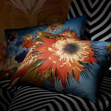 Surrearles'isme - Mosaique Decorative Pillow has a vibrant floral design, predominantly featuring orange and yellow flowers on a blue background, placed on a bed with a striped cover.