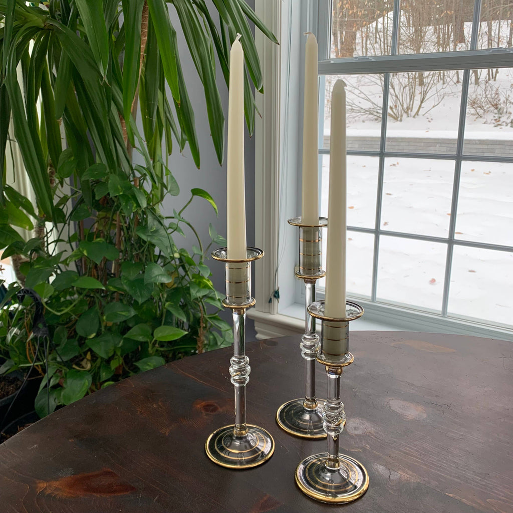 Three glass candlesticks in clear smoked glass with gold accents with candles in them on a wooden table near a window with a snowy view.
