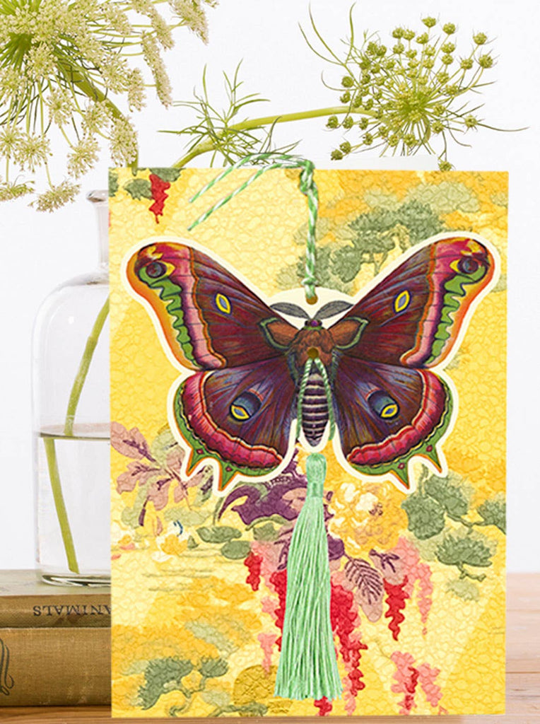Decorative card with a detachable colorful butterfly with a tassel on a yellow background, featuring greenery and books.