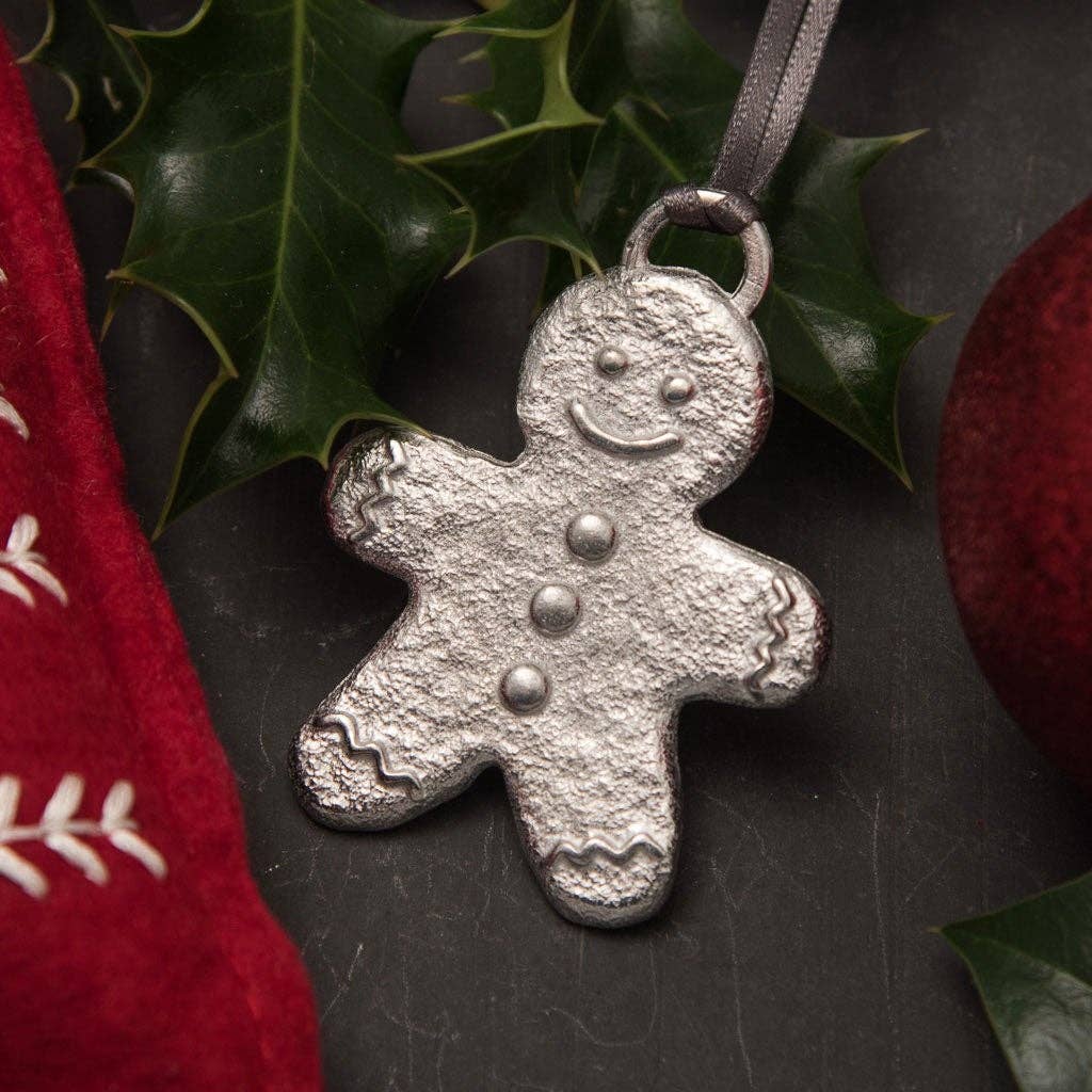 A pewter gingerbread man decoration with a happy facial expression, hanging from a ribbon and a metal chain, placed on a red textured surface with green leaves and a red and white checkered cloth in the background.