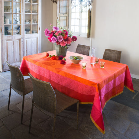 Parfums de bagatelle Nasturtium tablecloth woven with tone on tone flowers in bright orange and with pink and orange borders. On table in rustic yet elegant French dining room with wicker chairs and a glass chandelier, as well as flowers on the table.