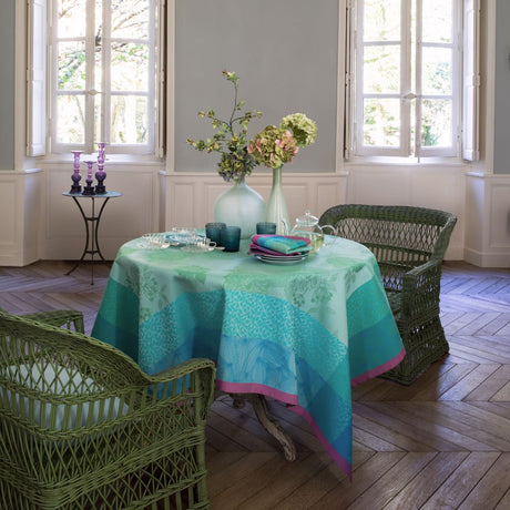 Table set with Parfums de Bagatelle Hydrangea tablecloth woven with tone on tone aqua flowers and a fuchsia border.  Woven rattan chairs, flowers on the table and open windows create an elegant relaxed setting.