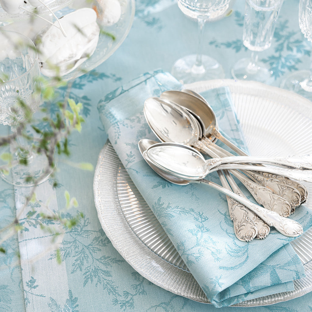 Jardin D'Eden Blue Napkin folded on glass plate with classic silver spoons piled on top.